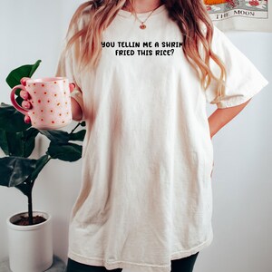 May include: Cream-colored oversized t-shirt with the text "YOU TELLIN ME A SHRIMP FRIED THIS RICE?" in black. The person is holding a pink mug with orange polka dots. The shirt is paired with dark green shorts.