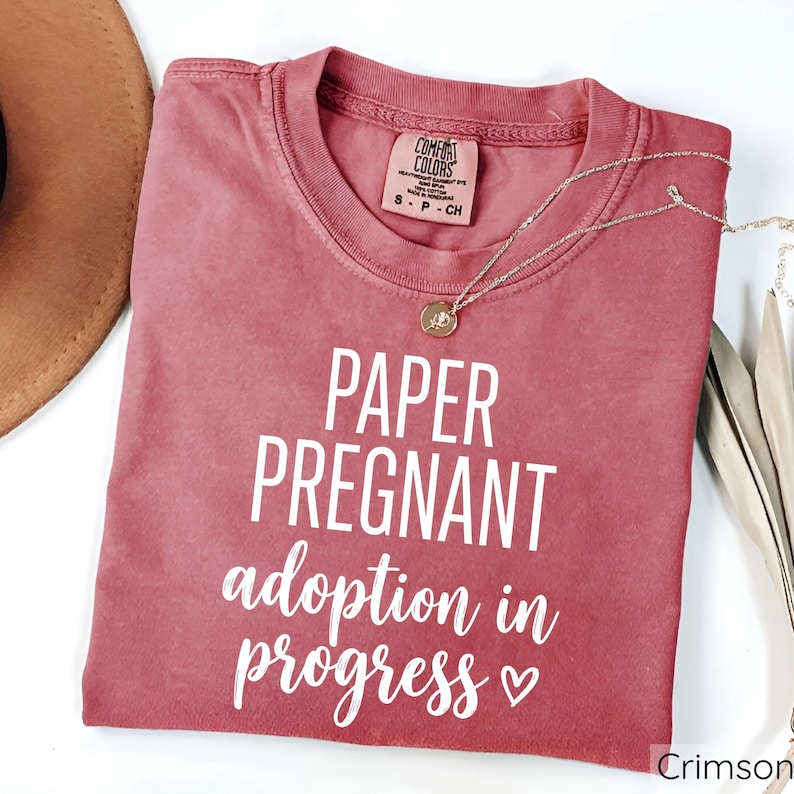 Pode incluir: Camiseta cor carmesim com o texto branco "PAPER PREGNANT adoption in progress". A camiseta tem gola redonda e mangas curtas. Um colar dourado com um pingente est&aacute; sobre a camiseta.