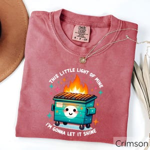 May include: A crimson red t-shirt with a graphic of a green dumpster on fire with the text "This little light of mine, I'm gonna let it shine".