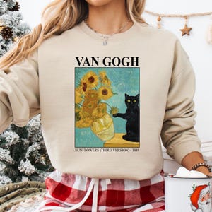 May include: Beige sweatshirt featuring a print of Van Gogh's "Sunflowers" painting with a black cat. The text "VAN GOGH" is above the image, and "SUNFLOWERS (THIRD VERSION) - 1888" is below. The sweatshirt has long sleeves.