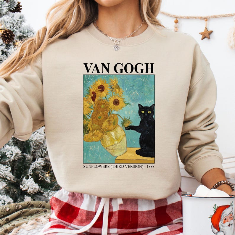 May include: Beige sweatshirt featuring a print of Van Gogh's "Sunflowers" painting with a black cat. The text "VAN GOGH" is above the image, and "SUNFLOWERS (THIRD VERSION) - 1888" is below. The sweatshirt has long sleeves.
