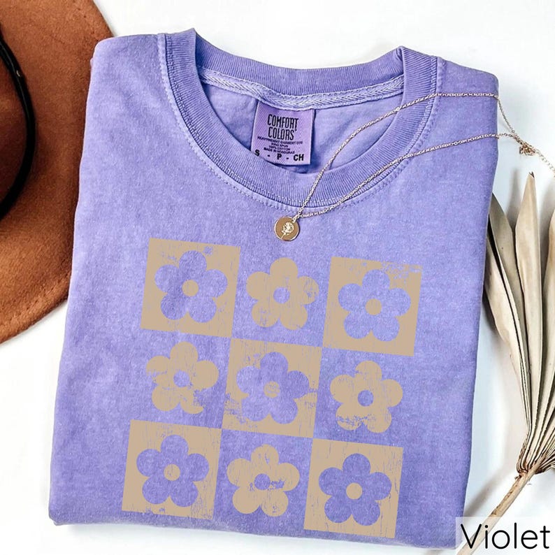 May include: A purple t-shirt featuring a retro-style design of brown flowers arranged in a grid pattern. The shirt has a "Comfort Colors" label on the front.