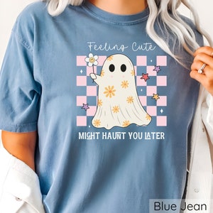 May include: Blue jean colored t-shirt with a graphic of a ghost holding a flower. The ghost has orange flower details and is set against a pink and white checkered background. Text reads "Feeling Cute Might Haunt You Later."