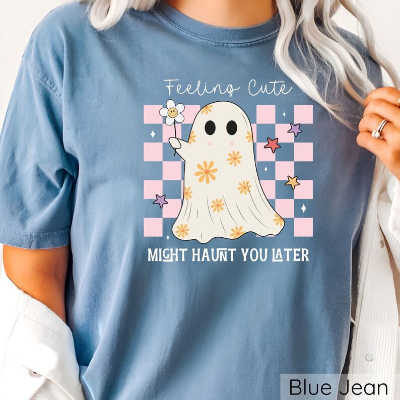 May include: Blue jean colored t-shirt with a graphic of a ghost holding a flower. The ghost has orange flower details and is set against a pink and white checkered background. Text reads "Feeling Cute Might Haunt You Later."