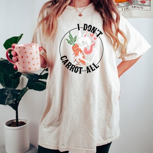 Może przedstawiać: Biały t-shirt z czarnym kołem graficznym przedstawiającym kresk&oacute;wkową marchewkę z czaszką i płomieniami. Wok&oacute;ł grafiki widnieje napis "I Don't Carrot All".
