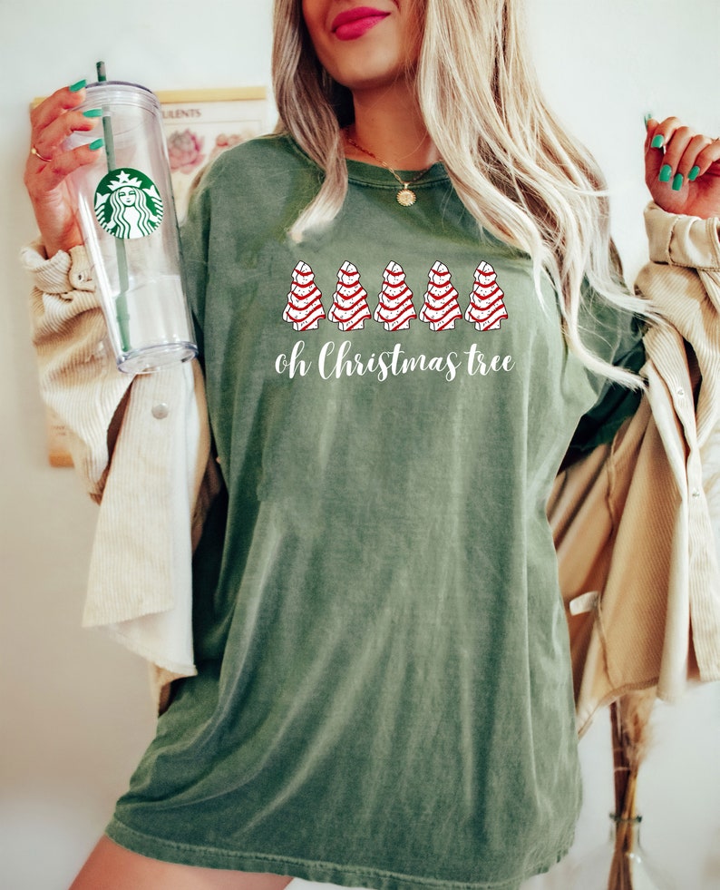 May include: Olive green t-shirt with the text "oh Christmas tree" and a graphic of six red and white striped Christmas trees. The shirt is a relaxed fit and has a vintage look.
