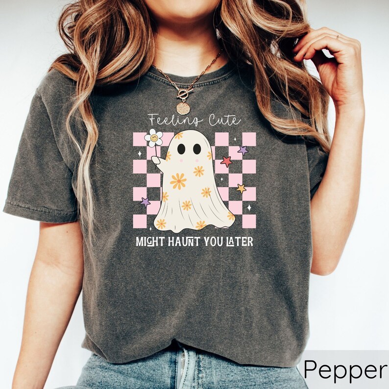 May include: Pepper-colored t-shirt featuring a ghost graphic holding a flower. The ghost is decorated with flowers and set against a pink and white checkered background. The text reads "Feeling Cute, Might Haunt You Later."