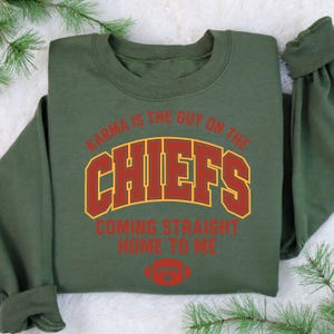 May include: An olive green sweatshirt with the text "Karma is the guy on the Chiefs coming straight home to me" in red and gold lettering. A red football graphic is below the text. The sweatshirt is folded.