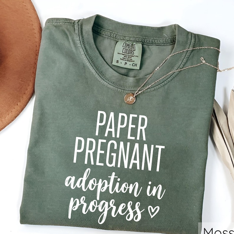 Pode incluir: Uma camiseta verde musgo com as palavras "PAPER PREGNANT adoption in progress" em escrita branca. Um colar de ouro com um pingente repousa sobre a camiseta. Um chap&eacute;u marrom &eacute; parcialmente vis&iacute;vel no canto superior esquerdo.