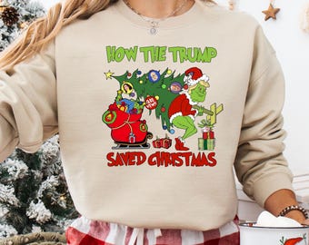May include: Beige sweatshirt with a Christmas-themed graphic. The design features a green Grinch-like character in a Santa hat, a Christmas tree, and the text "HOW THE TRUMP SAVED CHRISTMAS" in green and red.