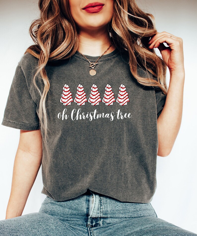 May include: Dark gray t-shirt featuring a festive design. The shirt displays five stylized Christmas trees with red and white stripes, above the words "oh Christmas tree" in a script font. The model is wearing blue jeans.