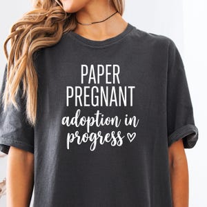 Pode incluir: Camiseta cinza escuro com o texto branco "PAPER PREGNANT adoption in progress". A camiseta tem mangas enroladas e gola redonda. O texto &eacute; em negrito e sem serifa.