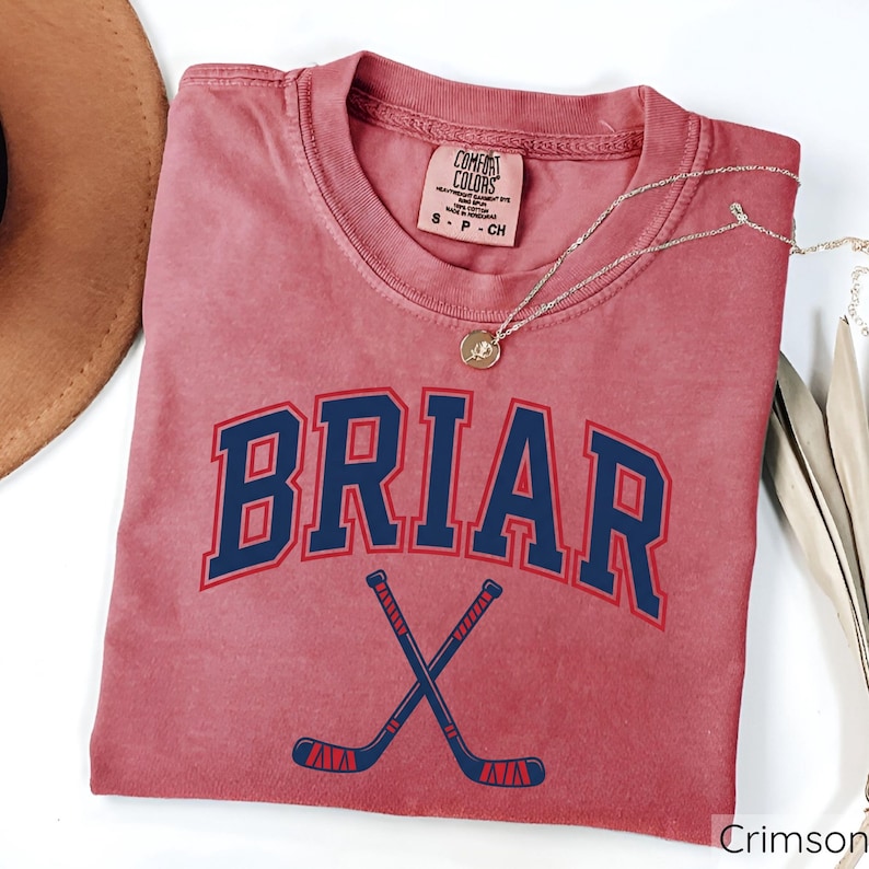 May include: A crimson short-sleeve t-shirt with a crew neckline. The front features the text BRIAR in dark blue with a red outline, arched above a graphic of two crossed dark blue hockey sticks with red accents. A Comfort Colors brand tag is visible.