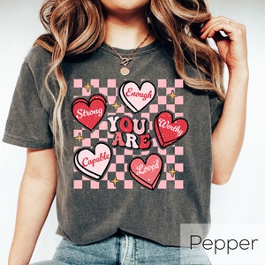 Puede incluir: Una camiseta gris con un fondo a cuadros rosa y blanco. Las palabras "You Are" est&aacute;n en el centro de la camiseta, rodeadas de corazones rosas con las palabras "Strong", "Enough", "Worthy", "Capable" y "Loved" en ellos.