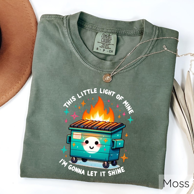 May include: A green t-shirt with a cartoon illustration of a dumpster on fire. The text "This little light of mine, I'm gonna let it shine" is printed on the shirt.