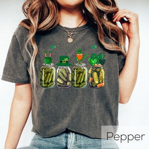 May include: A dark gray t-shirt with a graphic of four pickle jars with leprechauns on top. The leprechauns are wearing green hats and holding pots of gold. The text "Pepper" is visible in the bottom right corner of the image.