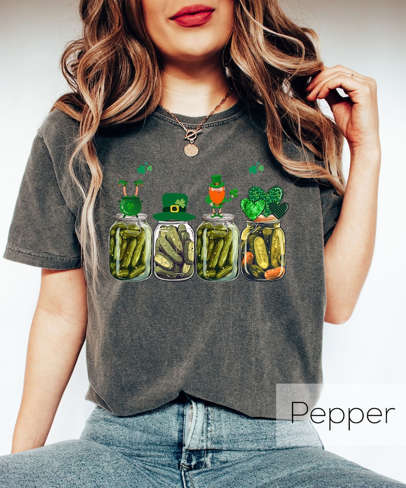 May include: A dark gray t-shirt with a graphic of four pickle jars with leprechauns on top. The leprechauns are wearing green hats and holding pots of gold. The text "Pepper" is visible in the bottom right corner of the image.