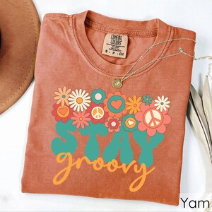 May include: A burnt orange t-shirt with a retro graphic design featuring flowers, peace signs, and the text "Stay groovy".