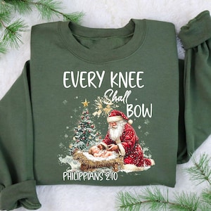 May include: Green sweatshirt with the text "EVERY KNEE Shall BOW" and "PHILIPPIANS 2:10". The design features Santa Claus kneeling beside a baby in a manger, with a Christmas tree in the background. A star and snowflakes are also in the design.