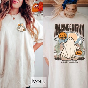 May include: Ivory-colored t-shirt with a Halloween-themed graphic. The front features a small design of a ghost with a pumpkin and cactus. The back displays a larger design with the text "Halloweentown Est. 1998" and a ghost holding a pumpkin balloon.