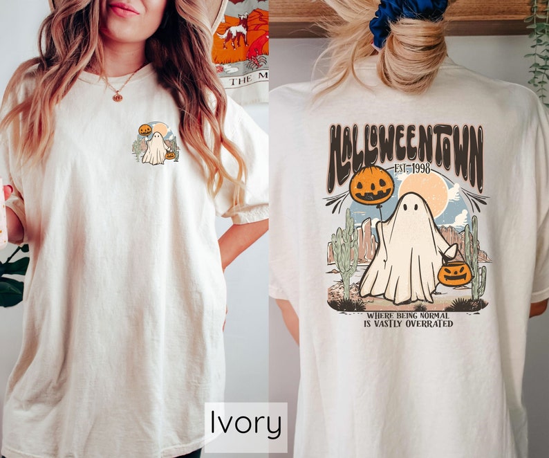 May include: Ivory-colored t-shirt with a Halloween-themed graphic. The front features a small design of a ghost with a pumpkin and cactus. The back displays a larger design with the text "Halloweentown Est. 1998" and a ghost holding a pumpkin balloon.