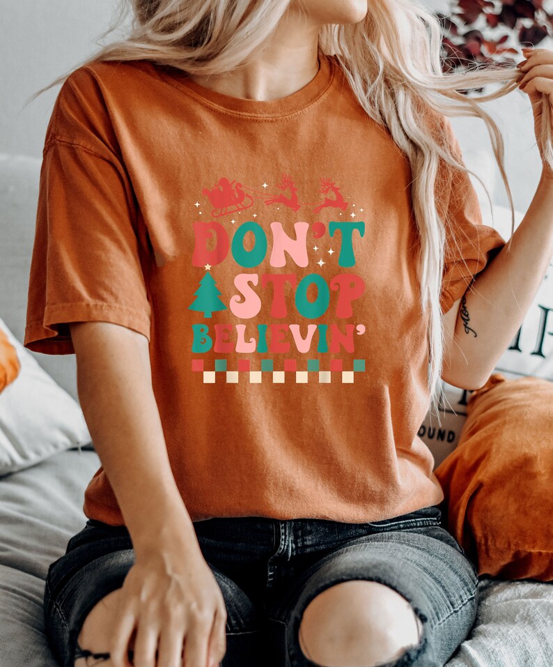 May include: A burnt orange short-sleeve t-shirt featuring a graphic print with the text "DON'T STOP BELIEVIN'" in a retro font. The design includes a silhouette of Santa in a sleigh with reindeer, a Christmas tree, and a checkered pattern at the base.