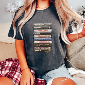 May include: Charcoal grey t-shirt displaying a graphic of stacked cassette tapes, including artists such as Madonna and George Michael. The design is printed on a casual tee, ideal for music enthusiasts and retro style.