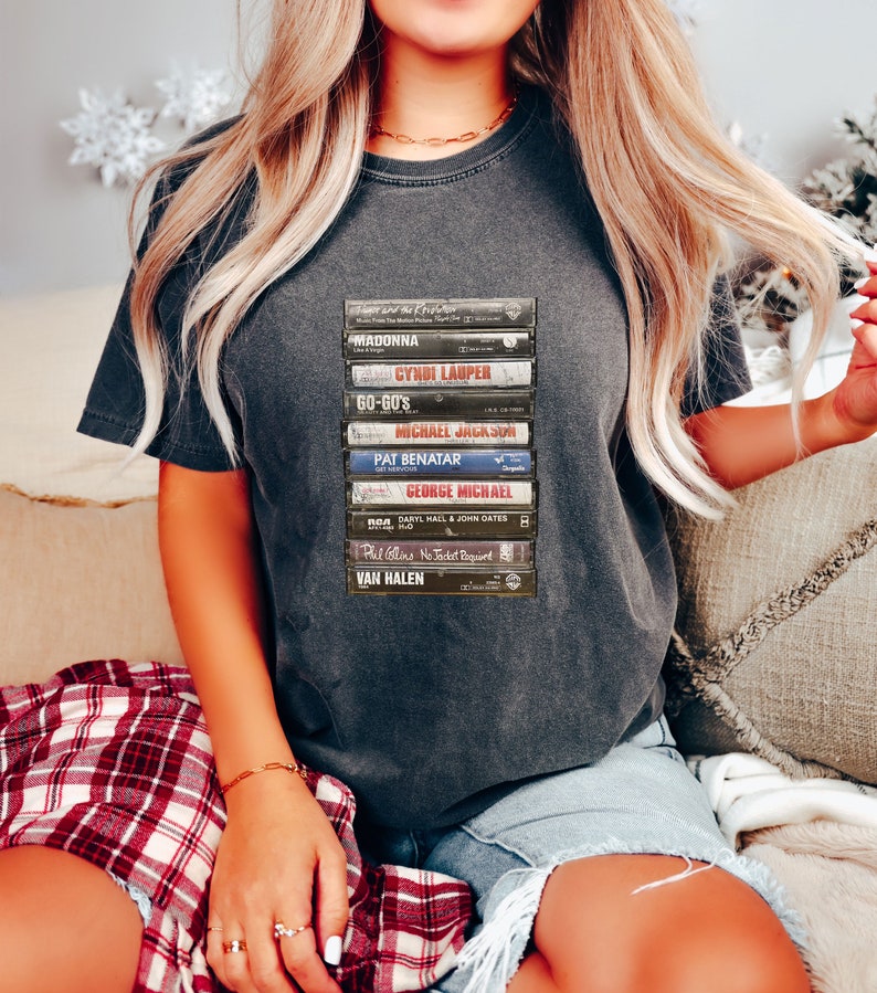 May include: Charcoal grey t-shirt displaying a graphic of stacked cassette tapes, including artists such as Madonna and George Michael. The design is printed on a casual tee, ideal for music enthusiasts and retro style.