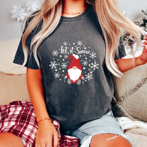 May include: A charcoal gray short-sleeve t-shirt featuring a white graphic of a gnome wearing a red hat, surrounded by white snowflakes and the text Let it Snow. The design is centered on the chest of the garment, which has a faded, vintage-style wash.