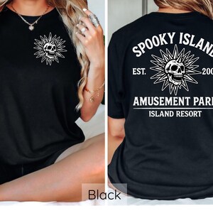 May include: Black t-shirt featuring a white skull design within a sunburst on the front and back. The back of the shirt displays the text "Spooky Island Amusement Park Island Resort" with the establishment year "2002".