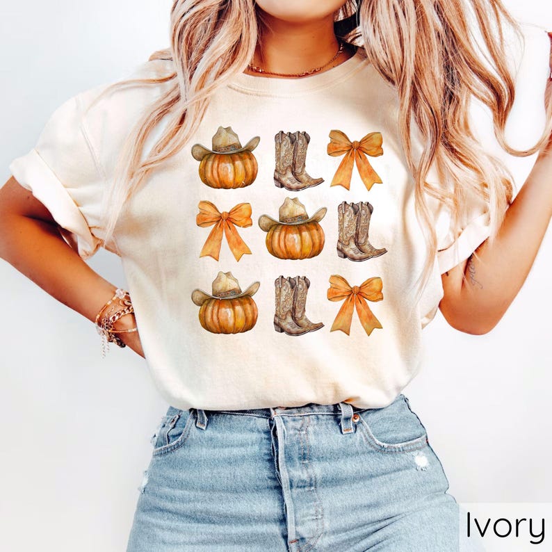 May include: Ivory-colored t-shirt featuring a repeating pattern of pumpkins with cowboy hats, cowboy boots, and orange bows. The design has a watercolor effect. The shirt is made of a soft material.