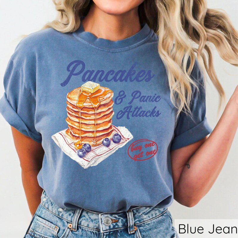 Op de afbeelding: Blauw jeans T-shirt met een afbeelding van een stapel pannenkoeken en de tekst "Pancakes & Panic Attacks" en "Buy one get one".