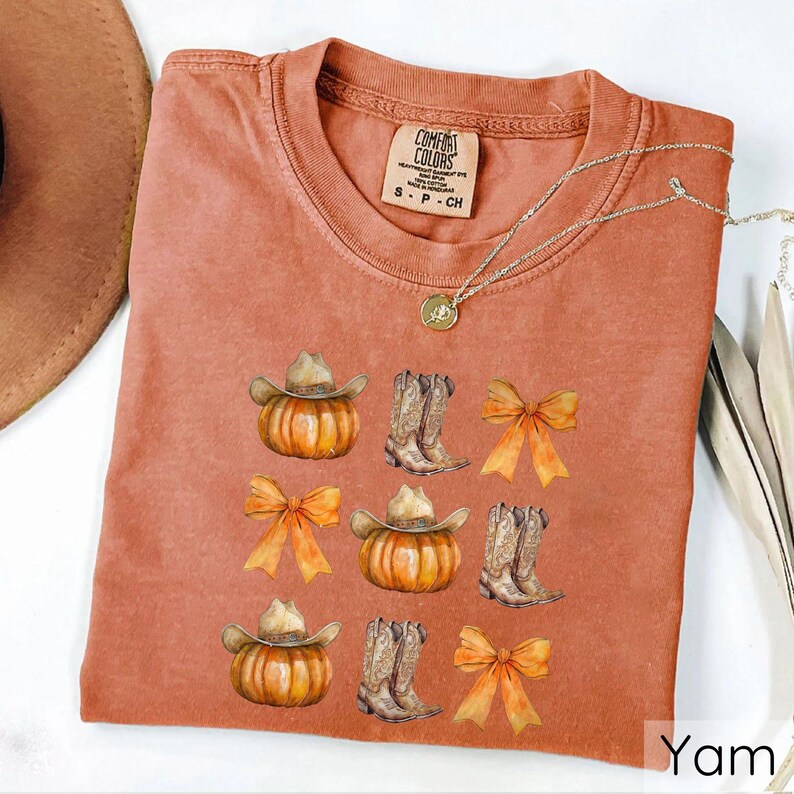 May include: A terracotta-colored t-shirt featuring a design of pumpkins wearing cowboy hats, cowboy boots, and orange bows. The shirt has a crew neck and short sleeves. The brand label "Comfort Colors" is visible at the top.