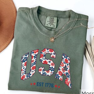 May include: A moss green t-shirt with the word "USA" in a floral pattern of red, white, and blue flowers. The text "EST 1776" is below the word "USA". A gold necklace with a pendant is on the shirt.