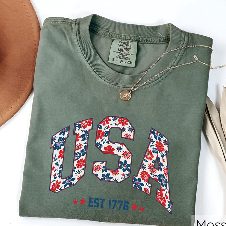 May include: A moss green t-shirt with the word "USA" in a floral pattern of red, white, and blue flowers. The text "EST 1776" is below the word "USA". A gold necklace with a pendant is on the shirt.
