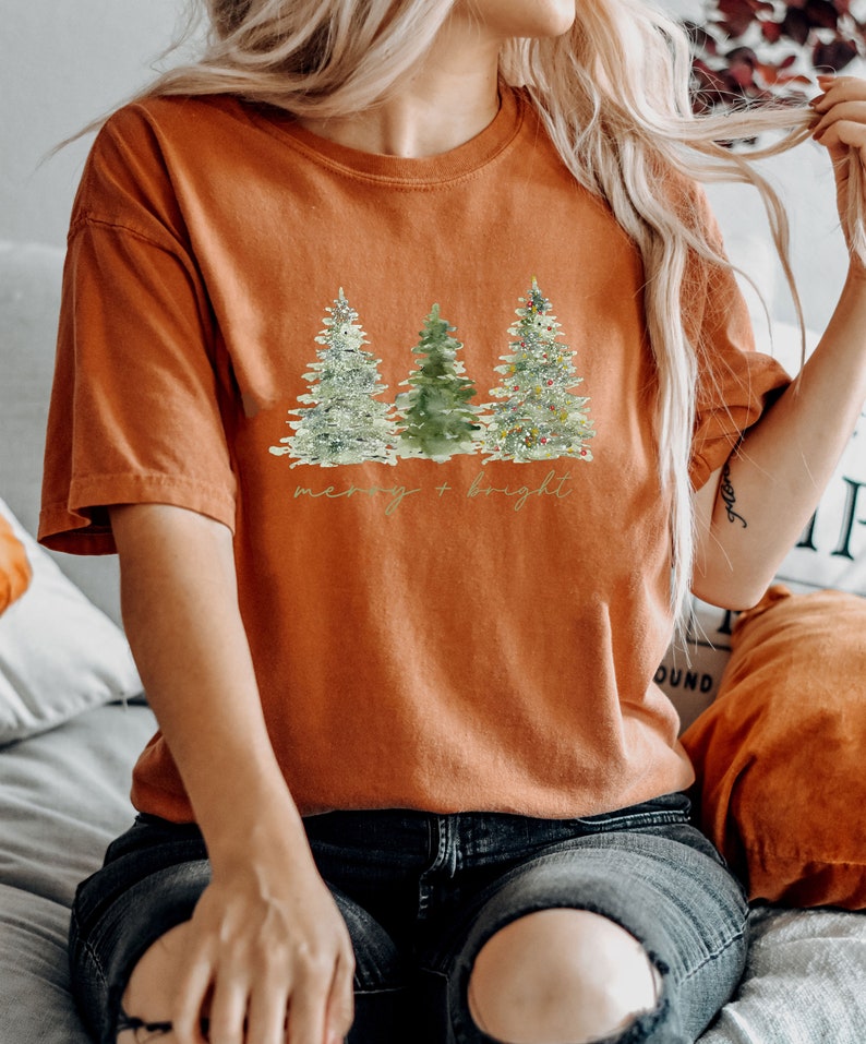 May include: An orange t-shirt featuring a watercolor design of three Christmas trees. The trees are in shades of green and white, with one decorated with colorful lights. The text "merry + bright" is written below the trees.