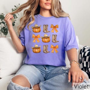 May include: A violet t-shirt featuring a repeating pattern of pumpkins wearing cowboy hats, cowboy boots, and orange bows. The word "Violet" is printed in white at the bottom right corner of the image.