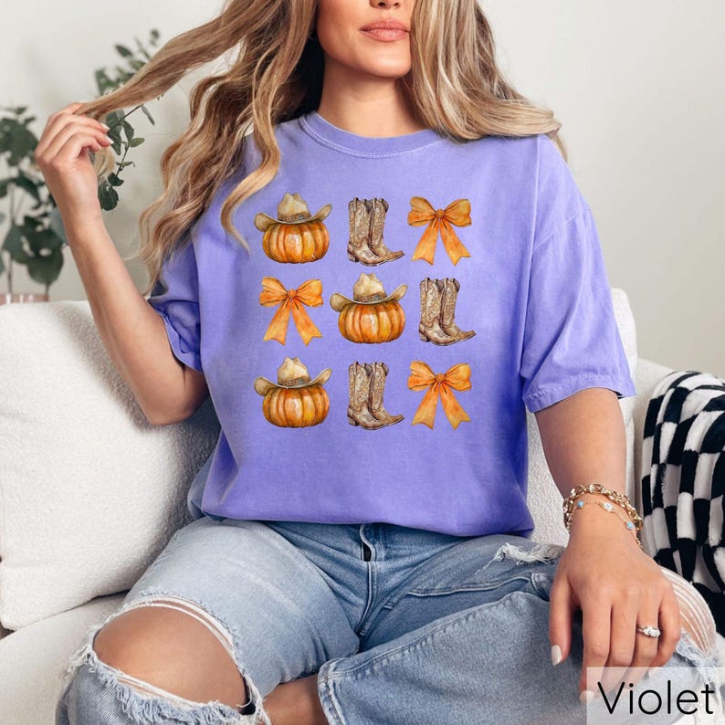 May include: A violet t-shirt featuring a repeating pattern of pumpkins wearing cowboy hats, cowboy boots, and orange bows. The word "Violet" is printed in white at the bottom right corner of the image.