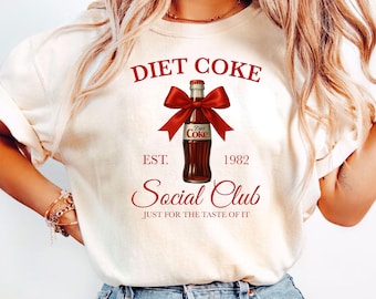 Può includere: T-shirt color crema con grafica di bottiglia di Diet Coke, fiocco rosso e la scritta "DIET COKE SOCIAL CLUB". Il design include "EST. 1982" e "JUST FOR THE TASTE OF IT" in rosso. La maglietta è abbinata a jeans blu.