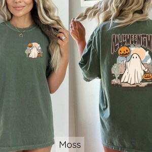 May include: Moss green t-shirt featuring a Halloween-themed graphic. The front graphic shows a ghost with a pumpkin and a desert scene. The back of the shirt displays the text "Halloweentown" with a ghost and pumpkins.