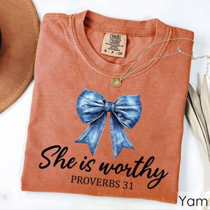 May include: A rust-colored t-shirt with a denim bow and the text "She is worthy Proverbs 31" printed on it.
