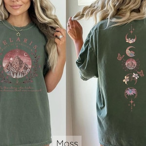 May include: A green t-shirt with a mountain scene and the text "Velaris City of Starlight" on the front. The back of the shirt features a vertical design of moons, flowers, and stars.
