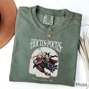 Puede incluir: Una camiseta verde musgo con las palabras "Hocus Pocus" en escritura blanca, con un gr&aacute;fico de tres brujas. La camiseta tambi&eacute;n tiene la frase "I Put a Spell on You" impresa debajo del gr&aacute;fico. Un collar de oro est&aacute; drapeado sobre la camiseta.