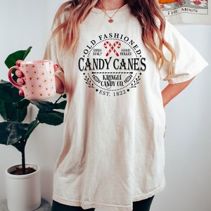 Pu&ograve; includere: T-shirt color crema con grafica circolare con la scritta "OLD FASHIONED CANDY CANES" e "KRINGLE CANDY CO. EST. 1822." Il design include due bastoncini di zucchero rossi e bianchi. La maglietta &egrave; indossata con pantaloncini verde scuro.