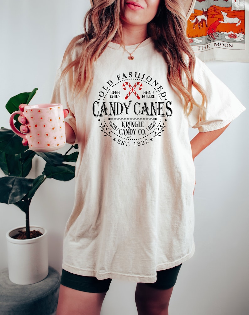 Pu&ograve; includere: T-shirt color crema con grafica circolare con la scritta "OLD FASHIONED CANDY CANES" e "KRINGLE CANDY CO. EST. 1822." Il design include due bastoncini di zucchero rossi e bianchi. La maglietta &egrave; indossata con pantaloncini verde scuro.