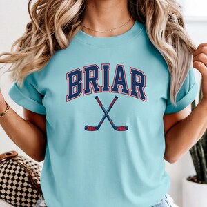 May include: A light blue t-shirt featuring the word BRIAR in a dark blue collegiate font with a red outline, positioned above a graphic of two crossed hockey sticks in dark blue with red accents on the handles and blades.