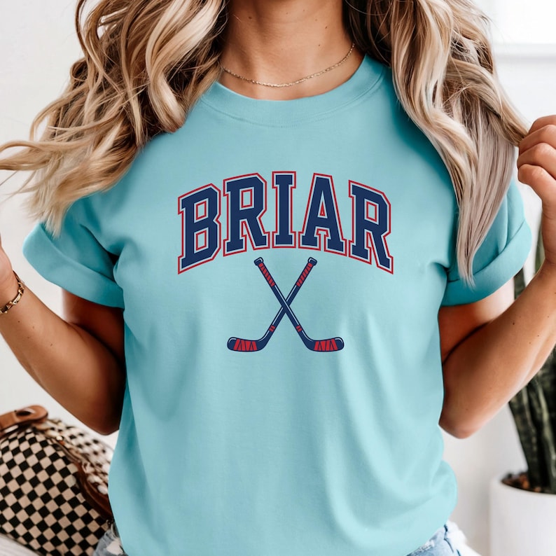 May include: A light blue t-shirt featuring the word BRIAR in a dark blue collegiate font with a red outline, positioned above a graphic of two crossed hockey sticks in dark blue with red accents on the handles and blades.