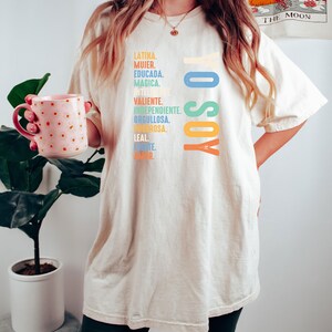 May include: A white oversized t-shirt with the text "Yo Soy" in rainbow colors. The text is surrounded by a list of Spanish words describing a woman's qualities.