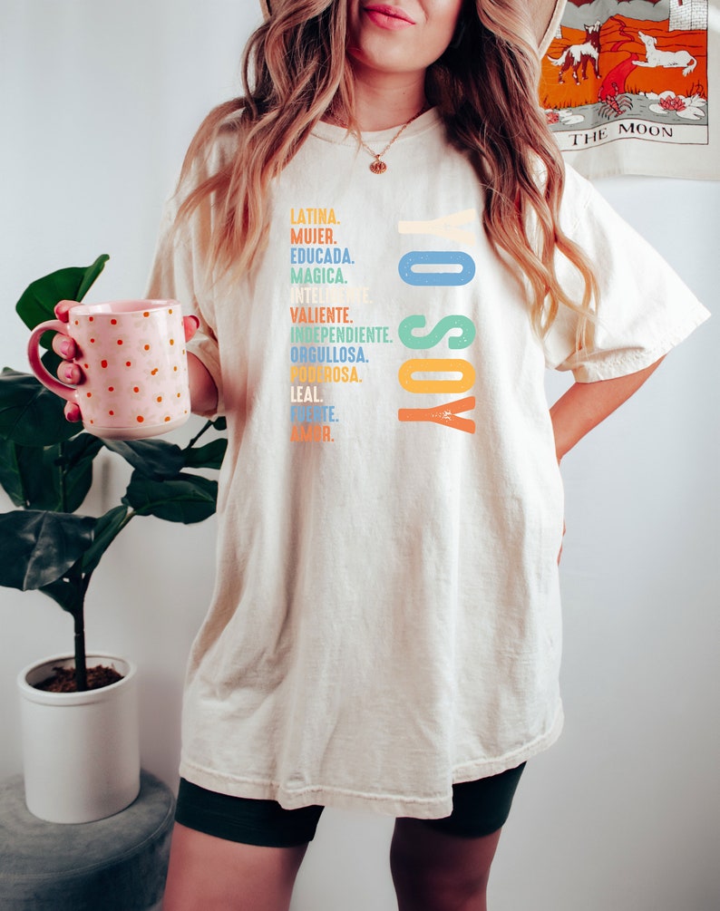 May include: A white oversized t-shirt with the text "Yo Soy" in rainbow colors. The text is surrounded by a list of Spanish words describing a woman's qualities.