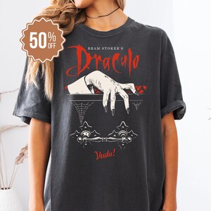 May include: Dark gray t-shirt featuring a graphic from Bram Stoker's Dracula. The design includes a reaching hand, spiderweb, and the word "Vudu!" in white. A brown 50% off sticker is in the upper left corner.